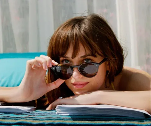 Woman with sunglasses lying on a towel, looking over papers, against a soft curtain backdrop.