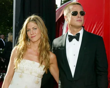 A woman in a white dress and a man in a tuxedo with sunglasses pose together on a red carpet.