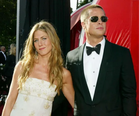 A woman in a white dress and a man in a tuxedo with sunglasses pose together on a red carpet.