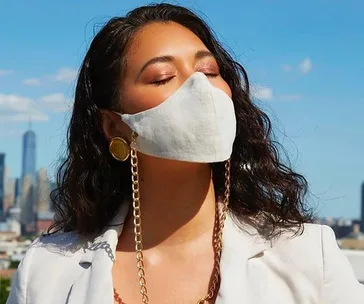 A woman in a white suit and mask stands against a clear blue sky, eyes closed, wearing gold earrings and mask chains.