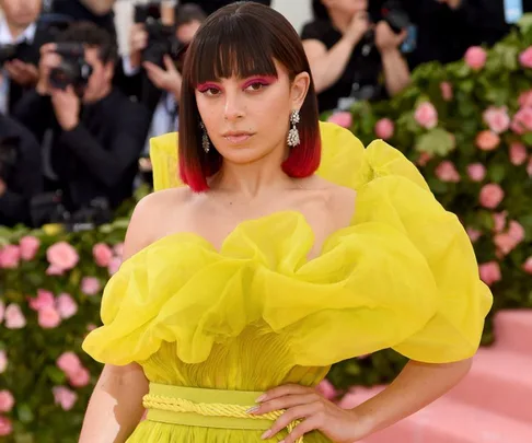 Woman in a vibrant yellow dress poses on a pink carpet with photographers and floral backdrop.