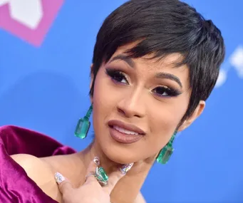 A woman with short hair and large green earrings poses on a blue and pink backdrop at an event.