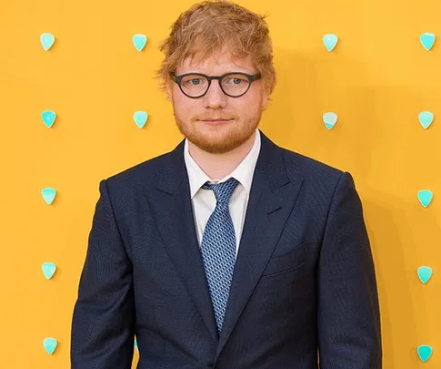 A man in a blue suit with glasses stands against a yellow background with teal dots.