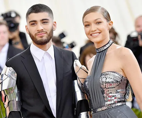 Gigi Hadid and Zayn Malik smiling at a formal event, with Gigi in a sequined gown and Zayn in a suit with metallic sleeves.