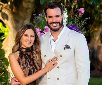 A couple smiling, standing outdoors, she in a brown dress, he in a beige suit, with flowers in the background.