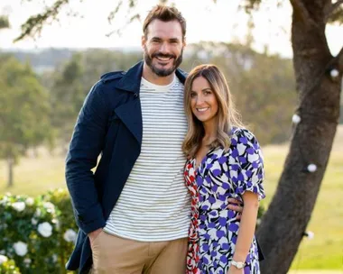 A couple posing together outdoors, smiling, with trees and a sunny background.