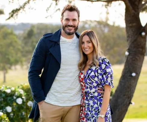 A couple posing together outdoors, smiling, with trees and a sunny background.