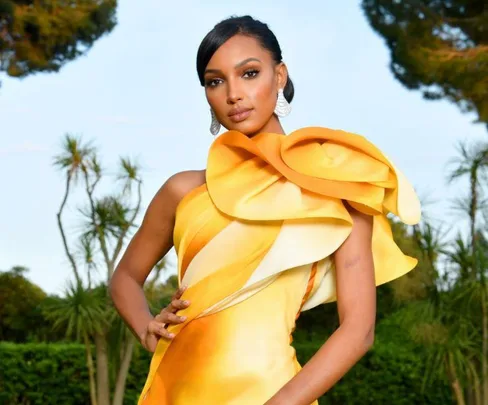 Woman in yellow-orange gown with ruffled shoulder posing outdoors near trees, wearing large earrings.