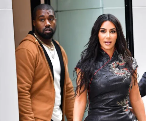 Kim Kardashian and Kanye West walking, Kanye wears a brown jacket, Kim in a black dress with dragon detail.