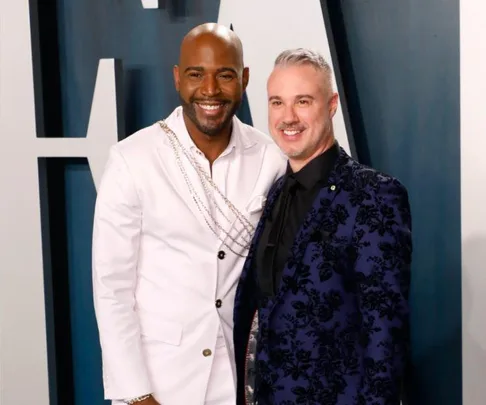 Two men smiling, one in a white suit with chains, the other in a blue floral suit, standing in front of a blue-gray wall.