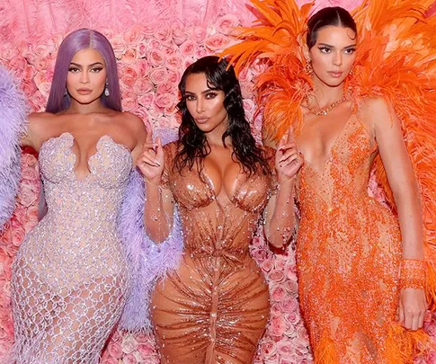 Three women stand before a pink floral backdrop, wearing glamorous, embellished evening gowns in purple, gold, and orange.