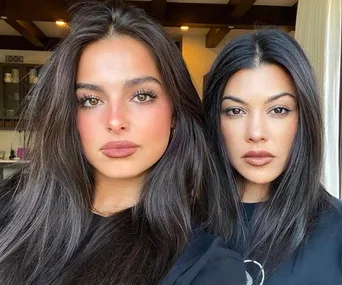 Two women with long, dark hair and serious expressions, standing side by side indoors.