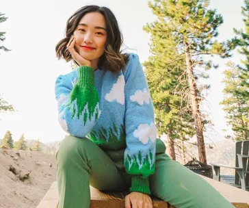 Woman in nature wearing a blue sweater with clouds and green pants, smiling in front of pine trees.