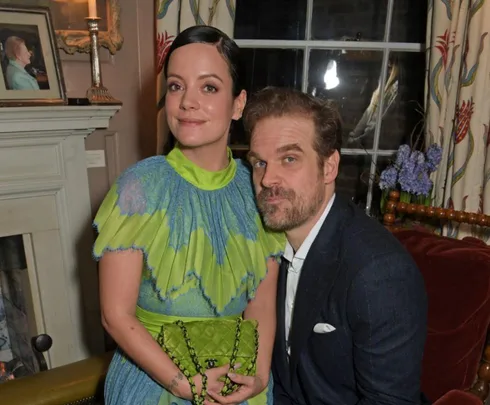 A couple dressed elegantly sitting together, with the woman holding a green purse, in a cozy living room setting.