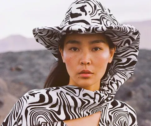 A person wearing a black and white marbled print hat and outfit, set against a rocky outdoor background.