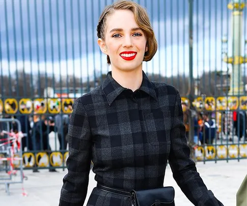 A person in a checkered dress with bright red lipstick smiling outdoors near a fence with a cloudy sky backdrop.