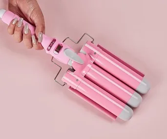 Hand holding a pink triple-barrel hair waver against a pink background.