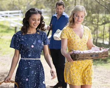 Two women walk outside, one in a blue dress and another in yellow carrying a picnic basket, followed by a man in a blue shirt.