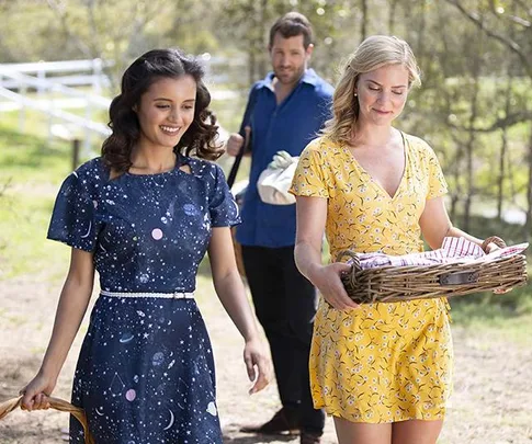 Two women walk outside, one in a blue dress and another in yellow carrying a picnic basket, followed by a man in a blue shirt.