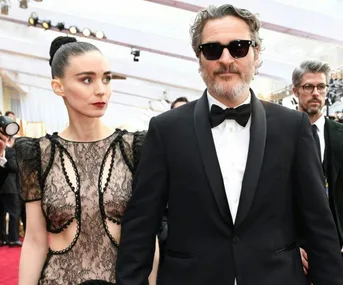 A couple on the red carpet; woman in lace dress, man in tuxedo with sunglasses.