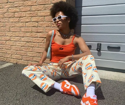 Person in orange top, logo pants, white sunglasses, seated against brick wall and garage door, wearing orange sneakers.