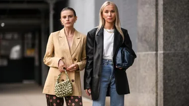 Two stylish women in blazers, one tan and one black, with a green bag and jeans, walking on a city street during NYFW 2020.