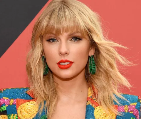 A woman with blonde hair and red lipstick wearing colorful clothing and green earrings, against a red background.