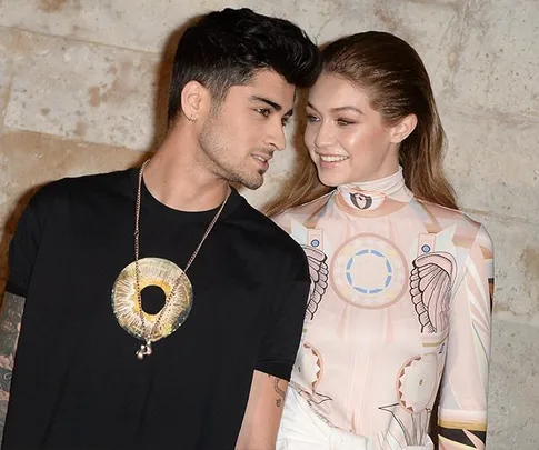 Zayn Malik and Gigi Hadid smiling at each other against a stone wall background.