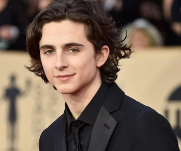 Young man with medium-length wavy hair in a black suit at a formal event, smiling subtly in front of a blurred background.