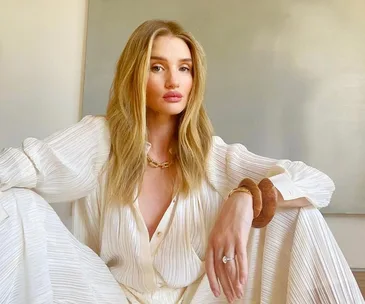 A person with long blonde hair in a white outfit sits indoors, looking at the camera.