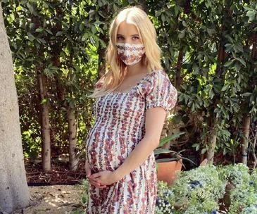 Pregnant woman in floral dress with matching mask stands in garden, cradling her belly.