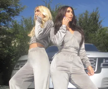 Two women in matching grey tracksuits pose confidently in front of a car, one with long blonde hair, the other with dark hair.