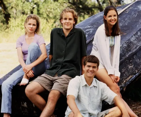 Four young adults from "Dawson's Creek" sitting outdoors, smiling, with trees in the background.