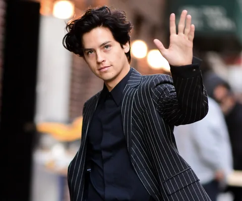 A person in a pinstripe suit waves with a neutral expression on a city street.