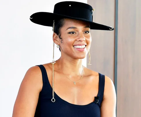 Alicia Keys smiling, wearing a black hat and tank top, with chain earrings and a delicate necklace.