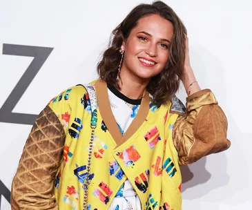 Alicia Vikander smiling at an event, wearing a colorful patterned yellow jacket.