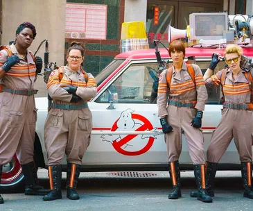 Four women in Ghostbusters uniforms stand confidently in front of the iconic Ecto-1 car.