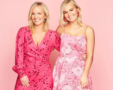 Two women smiling, wearing vibrant pink dresses with heart and floral patterns, posing against a pink background.