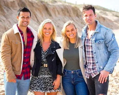 A group of four smiling people standing together on a beach, dressed in casual and denim outfits.