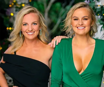 Two women smiling, one wearing a black dress, the other in a green dress, with a blurred, festive background.