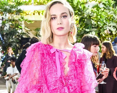 Blonde woman in a pink ruffled dress at an outdoor event, surrounded by people and greenery.