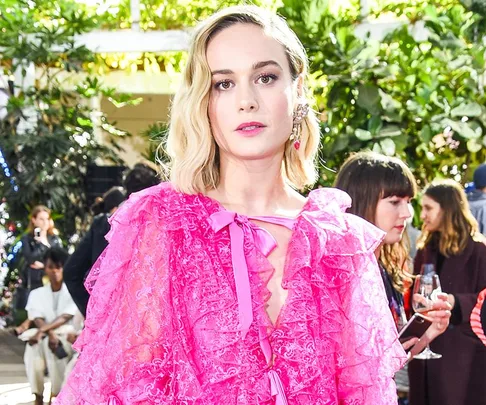 Blonde woman in a pink ruffled dress at an outdoor event, surrounded by people and greenery.