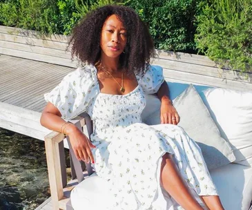 Woman in a floral dress seated outdoors on a white cushion by greenery.