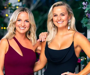 Two women with blonde hair smiling, wearing elegant evening dresses and large earrings, standing in a floral-decor setting.