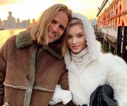 A couple in winter clothing smiles by the waterfront at sunset.