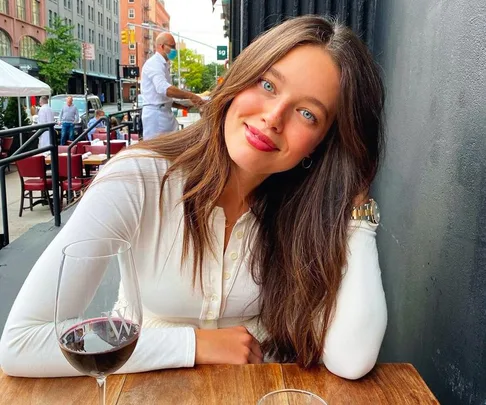 Woman with long brown hair in a white top smiles at a cafe table with a glass of red wine.