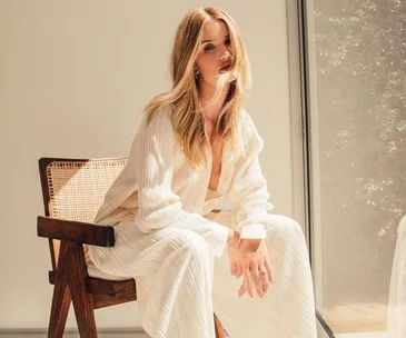 Woman in a stylish white outfit sitting on a wooden chair by a window with sunlight streaming in.