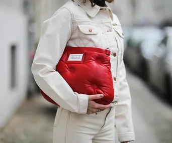 Person in beige outfit holding red quilted bag with label, standing on blurred city street.