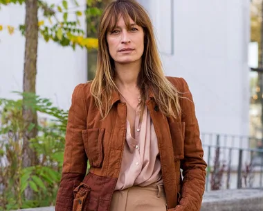 A woman with long hair wearing a brown jacket and beige shirt stands outside with blurred greenery in the background.