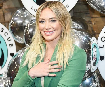 Blonde woman in a green jacket smiles at an event with reflective balloons in the background.